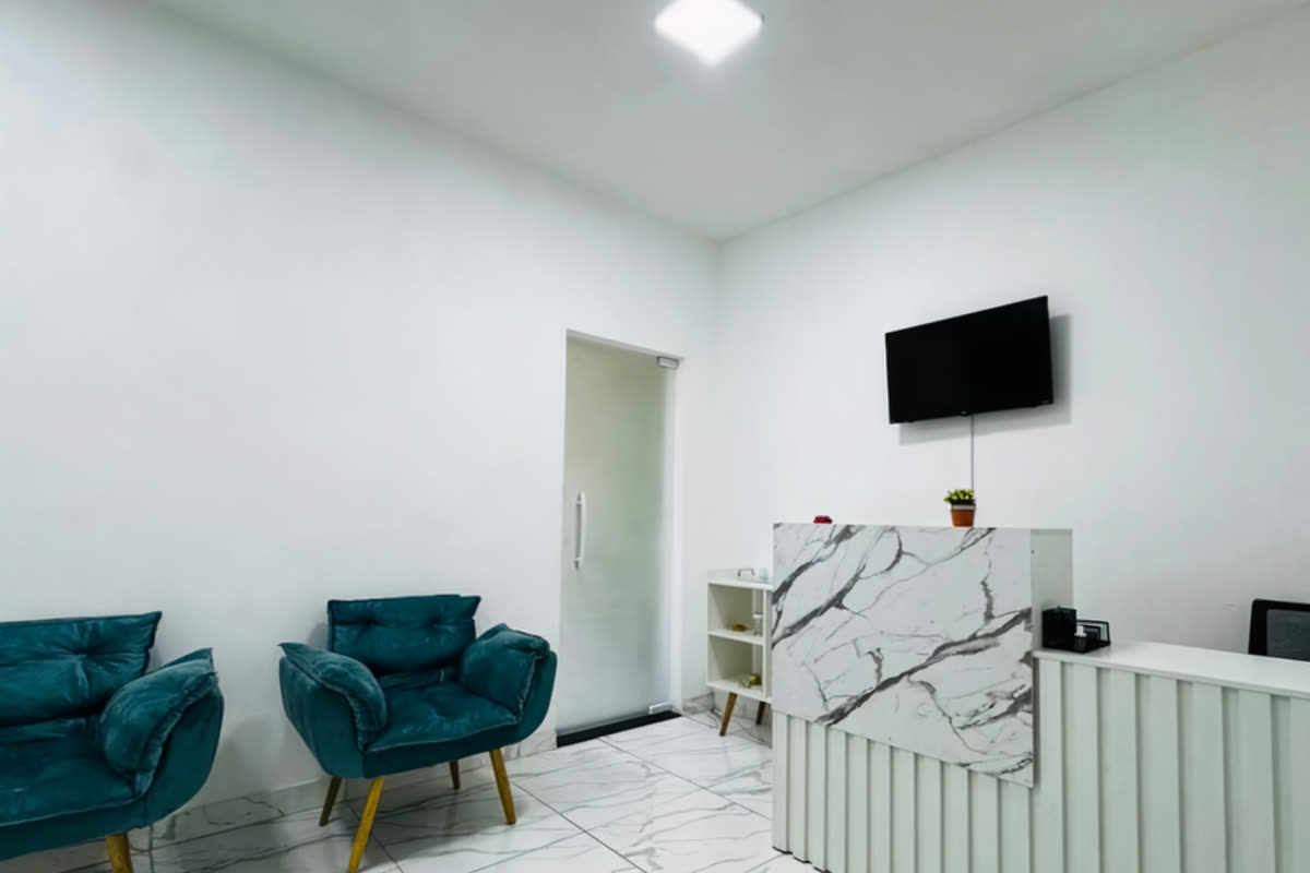 Recepção da Sorrir Bem, com poltronas de veludo azul-petróleo, piso de porcelanato marmorizado, balcão branco e televisão na parede